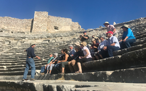 People sitting an greek ruins listening to a tour guide